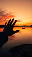 Silhouette of human hand reaching toward vibrant orange sunset over calm lake with distant boat, dramatic sky reflection on water surface.