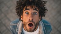 Shocked young mixed race man with curly hair and wide eyes expressing surprise and disbelief with open mouth against blurred background for advertising campaigns.