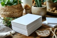 Stack of blank white paper cards on wooden table with green plants, woven basket, and ceramic decorative elements creating cozy home office atmosphere.