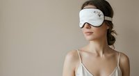 Young Caucasian woman wearing white sleep mask with star pattern relaxing in silk camisole against neutral beige background for wellness and self-care concepts.