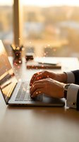 Professional businessman hands typing on laptop with glowing digital particles and warm sunlight streaming through office window creating productive workspace atmosphere.