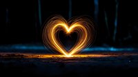 Glowing heart shape created with light painting technique against dark forest background, illuminating ground with warm golden light trails. Long exposure photography.