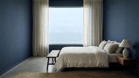 Serene bedroom with ocean view, neutral bedding, blue walls, and flowing curtains creating a calm, minimalist coastal retreat space.