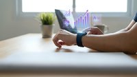 Man wearing smartwatch displaying holographic business analytics and financial growth charts at desk with laptop and plant, representing digital technology and data visualization for business intelligence applications.