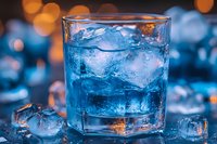 Refreshing blue cocktail with ice cubes in glass against blurred bokeh background, close-up view. Vibrant drink photography with cool tones and orange highlights.