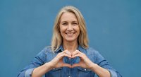 Smiling blonde middle-aged Caucasian woman making heart shape with hands wearing denim shirt against blue background for wellness and love campaigns.