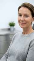 Confident middle-aged Caucasian woman in gray sweater smiling gently in a modern clinic or office interior setting.