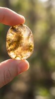 Hand holding translucent amber gemstone with natural inclusions against sunlight creating warm golden glow and bokeh background for jewelry design.