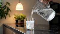 Water being poured into a glass on a modern kitchen counter with pendant lighting and potted plants, suitable for beverage, hydration and lifestyle content.