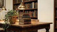 Stacked vintage books with brass lamp and potted plant on wooden desk against bookshelf background for interior design and home decor projects.