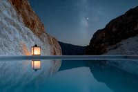Glowing lantern reflects in still mountain lake water at night, with dramatic canyon walls and starry sky creating serene wilderness atmosphere.