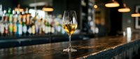 Glass of white wine on wooden bar counter in upscale restaurant with blurred bottles and warm lighting creating elegant dining atmosphere.