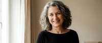 Happy mature woman with gray curly hair smiling warmly in black top at home, representing confidence and positive aging lifestyle.