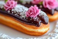 Chocolate glazed eclair decorated with pink sugar rose and silver sprinkles on white plate, close up view of French pastry dessert with dark ganache coating.