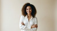 Professional African American businesswoman with curly hair smiling confidently with crossed arms in white shirt against neutral background for corporate use.