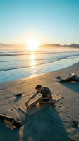 Young man stretching on a mat at a sandy beach during golden sunrise, with ocean waves, misty mountains, and driftwood in the background.