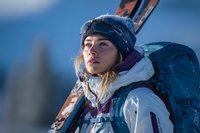 Young Caucasian woman in winter sports gear carrying skis on mountain with backpack, wearing knit hat and looking at horizon during adventure.