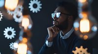 Young African American businessman in sunglasses thinking thoughtfully against glowing cog wheel background with bokeh lights, representing innovation and strategic planning.