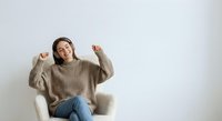 Young woman in beige sweater celebrating success with raised fists while sitting in white armchair against neutral background for lifestyle content.