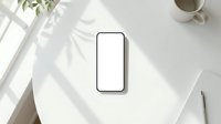 Smartphone with blank white screen on clean white desk with natural sunlight shadows, coffee cup and plant for mockup presentations and app development.