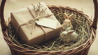 Rustic gift box wrapped in kraft paper with twine bow nestled in wicker basket with straw and small bunny figurine for Easter celebration.