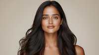 Young woman with glowing bronzed skin, long dark wavy hair, and natural makeup smiling confidently against neutral beige background. Professional beauty and cosmetics portrait.