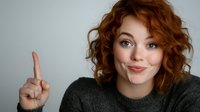Young woman with curly red hair and bright blue eyes pointing finger up showing attention gesture, wearing casual sweater against light background.