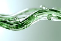 Clear water drops on flowing green glass wave formation against light background, creating dynamic abstract composition with transparent spheres and curves.