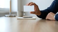 Adult male with medium skin tone wearing dark business attire, holding blank white card at wooden desk with laptop and coffee cup, ideal for corporate branding and business communications.