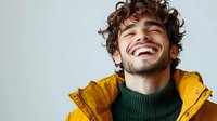 Young man with curly brown hair wearing yellow winter jacket and green turtleneck sweater laughing with genuine joy and happiness against light background.