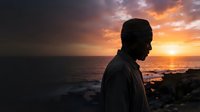Silhouette of a man in profile standing on rocky coastline at sunset, gazing toward ocean horizon with dramatic golden and orange sky, contemplative mood for meditation and lifestyle concepts.