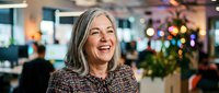 Middle-aged woman with long gray hair smiling in modern office lobby with holiday decorations and blurred colleagues in background.