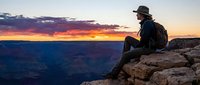 Hiker in dark clothing and hat sitting on rocky cliff edge overlooking vast canyon landscape during dramatic sunset with purple and orange sky.