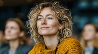 Professional blonde woman with curly hair in orange sweater smiling confidently during business conference or seminar with colleagues in background.