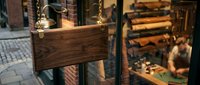 Wooden blank sign hanging on glass door of artisan leather workshop with craftsman working in background.