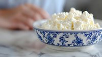 Fresh cottage cheese in traditional blue and white porcelain bowl on marble surface, with blurred hands in background, natural daylight setting.