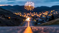 Glowing firework bursts over mountain valley at dusk, with bokeh lights from street lamps creating magical atmosphere along illuminated road stretching into distance.