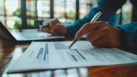 Business professional analyzing financial data and charts on paper documents while working at wooden desk in modern office environment.