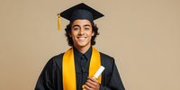 Young Hispanic male graduate in black cap and gown with yellow stole holding diploma, smiling against beige background.