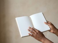Open book with blank white pages held by hands of a person with dark skin tone against neutral beige background, suitable for publishing, education, literature, and branding mockups.