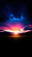 Dramatic mountain sunset with vibrant pink and blue sky, golden sunburst breaking through dark clouds over silhouetted peaks and meadow landscape.
