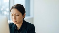 Young Asian businesswoman experiencing stress and headache while working at modern office desk near window during daytime.