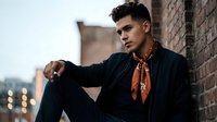 Young handsome Hispanic man in black suit with orange scarf sitting against brick wall in urban setting during golden hour for fashion and lifestyle concepts.