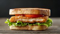 Crispy chicken sandwich with fresh lettuce and tomato on toasted bread, served on rustic wooden surface against dark background. Close up food photography.