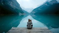 Woman sitting on wooden dock facing turquoise alpine lake surrounded by forested mountains, contemplative travel and nature exploration scene.