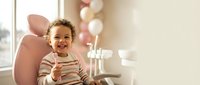 Happy mixed-race toddler girl sitting in pink chair at birthday party with balloons, celebrating special occasion indoors with natural window light.