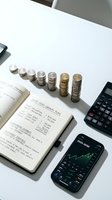 Financial planning workspace with stacked coins, calculator, notebook and smartphone showing investment charts on white desk for budgeting and money management.