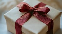 Elegant gift box wrapped in cream paper with burgundy red ribbon bow, soft focus close-up view showing texture and festive presentation for special occasions.