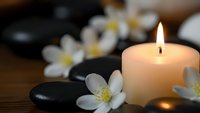 Spa wellness scene with burning candle, black massage stones and white flowers on wooden surface for relaxation and meditation therapy.