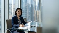 Professional Asian businesswoman in navy suit working at modern office desk with city skyline view through windows for corporate presentations.